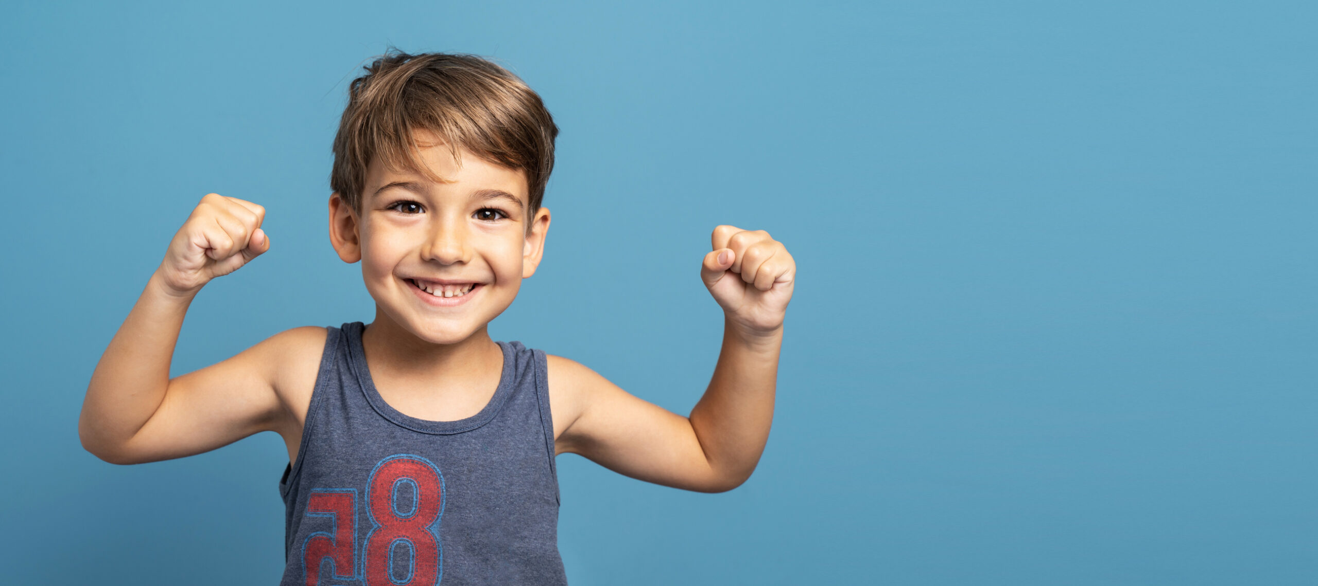 Front view of small caucasian boy four years old standing in front of blue background studio shot standing confident flexing muscles smiling growing up and strength and health concept